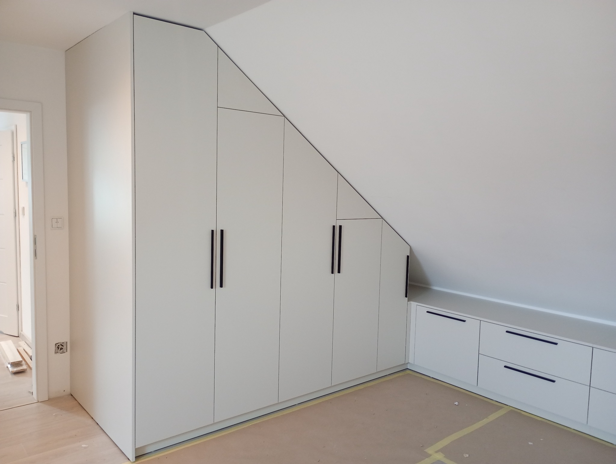 Biała szafa wnękowa z czarnymi uchwytami, dopasowana do skosu poddasza. Widoczne listwy przypodłogowe i fragment podłogi. Minimalistyczny design.