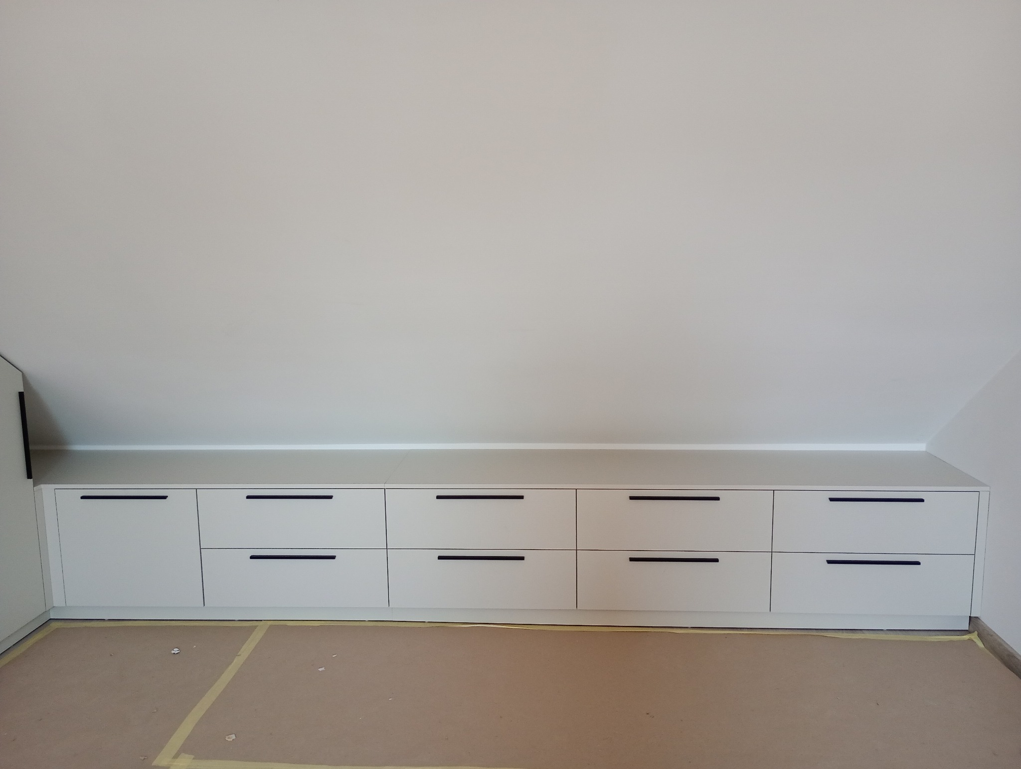 Biała zabudowa meblowa ze szufladami pod skosem dachu, z minimalistycznymi czarnymi uchwytami, na podłodze zabezpieczonej kartonem.