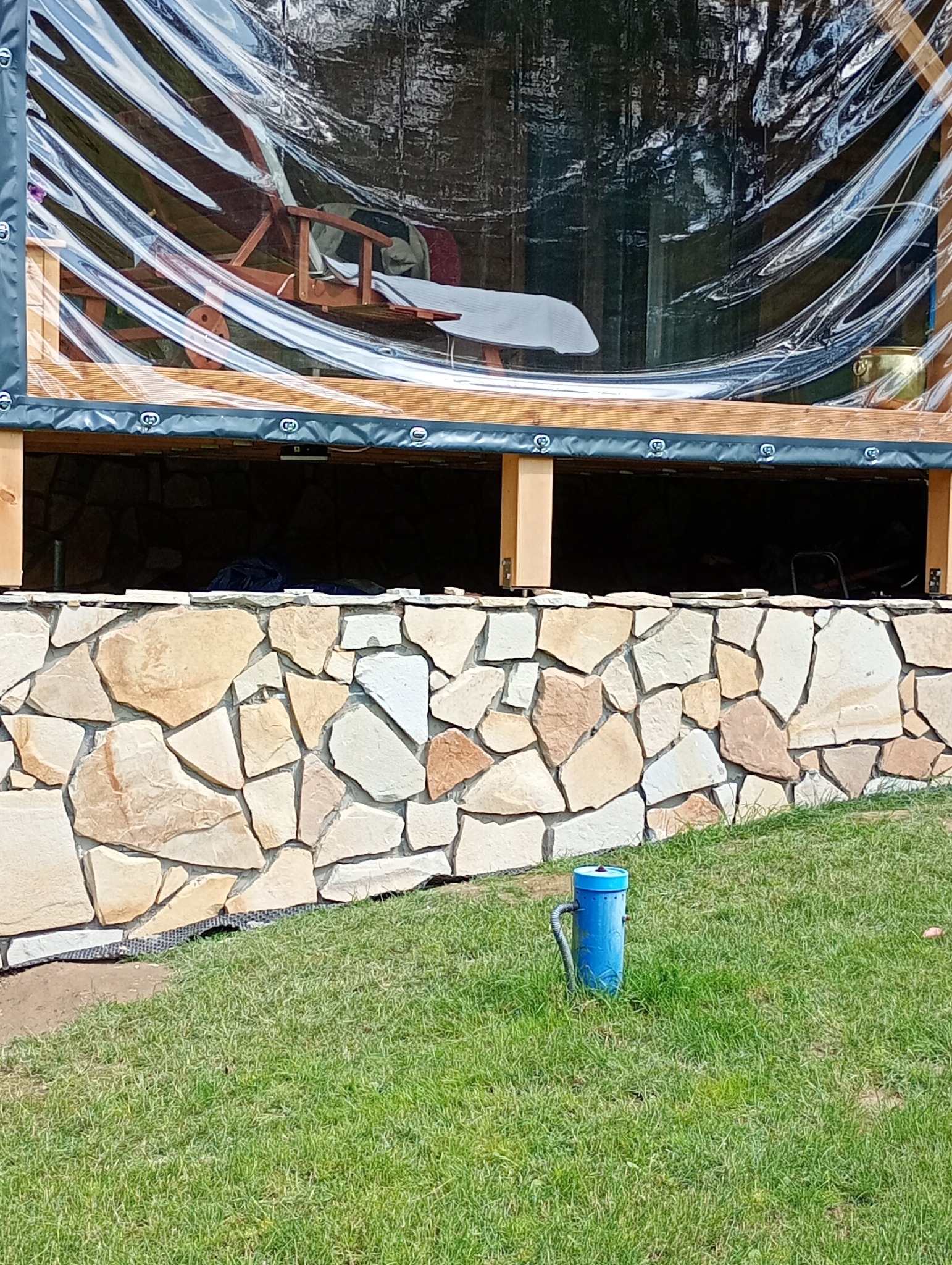 Zadaszony taras z drewnianą podłogą, osłonięty folią i kamienną podmurówką. Na tarasie widoczny fotel. Na trawniku niebieski hydrant.