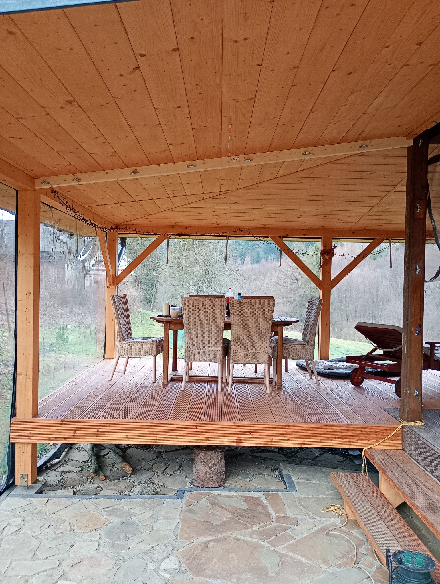 Drewniany taras z zadaszeniem i meblami ogrodowymi, widok na las. Zastosowano deski tarasowe z wyraźnym usłojeniem. Zadaszenie wykonane z drewna, z elementami dekoracyjnymi.