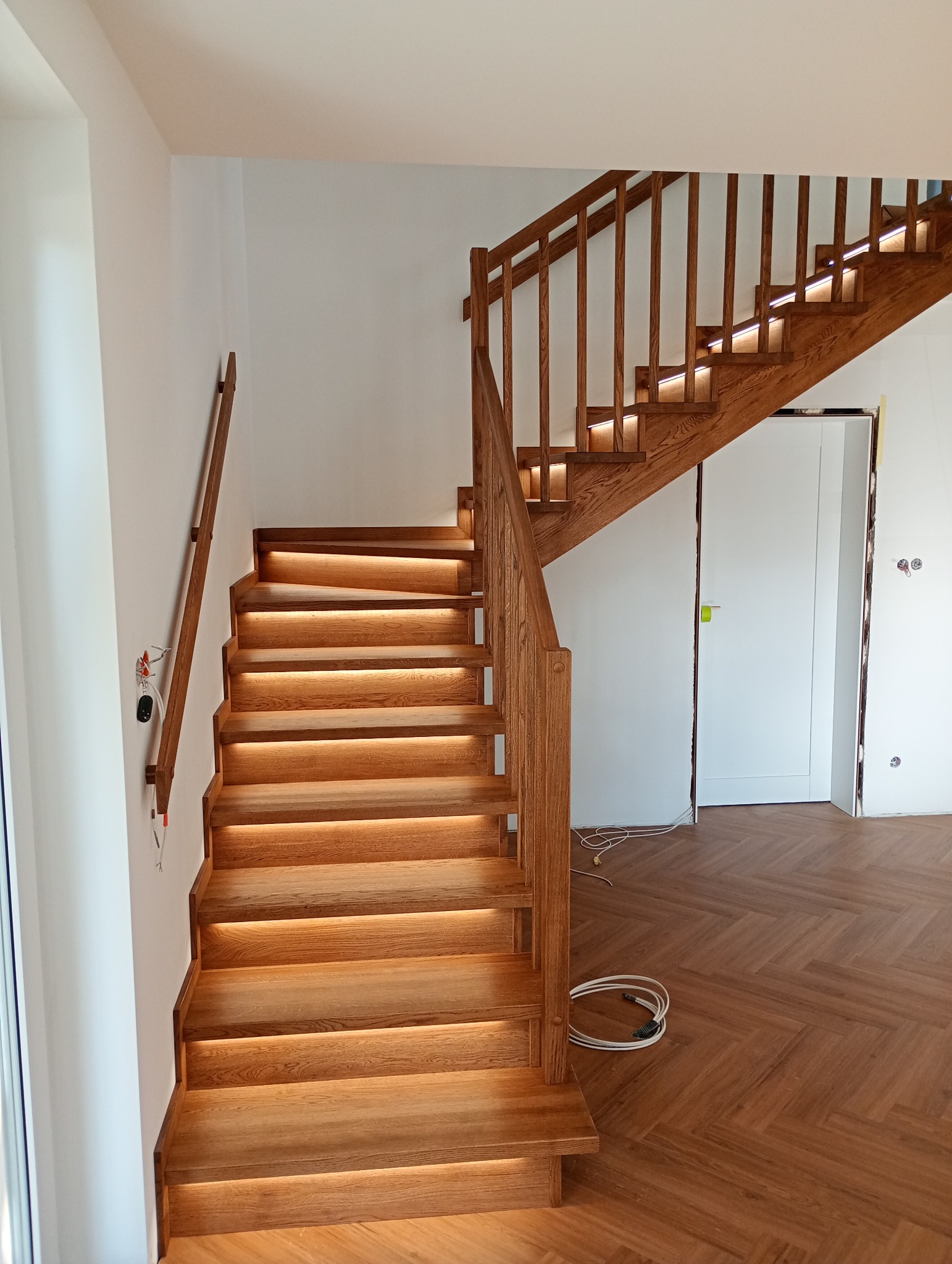 Drewniane schody z podświetleniem LED, dębowe stopnie i balustrada, parkiet w jodełkę na podłodze, białe ściany. Nowoczesne wykończenie wnętrza.