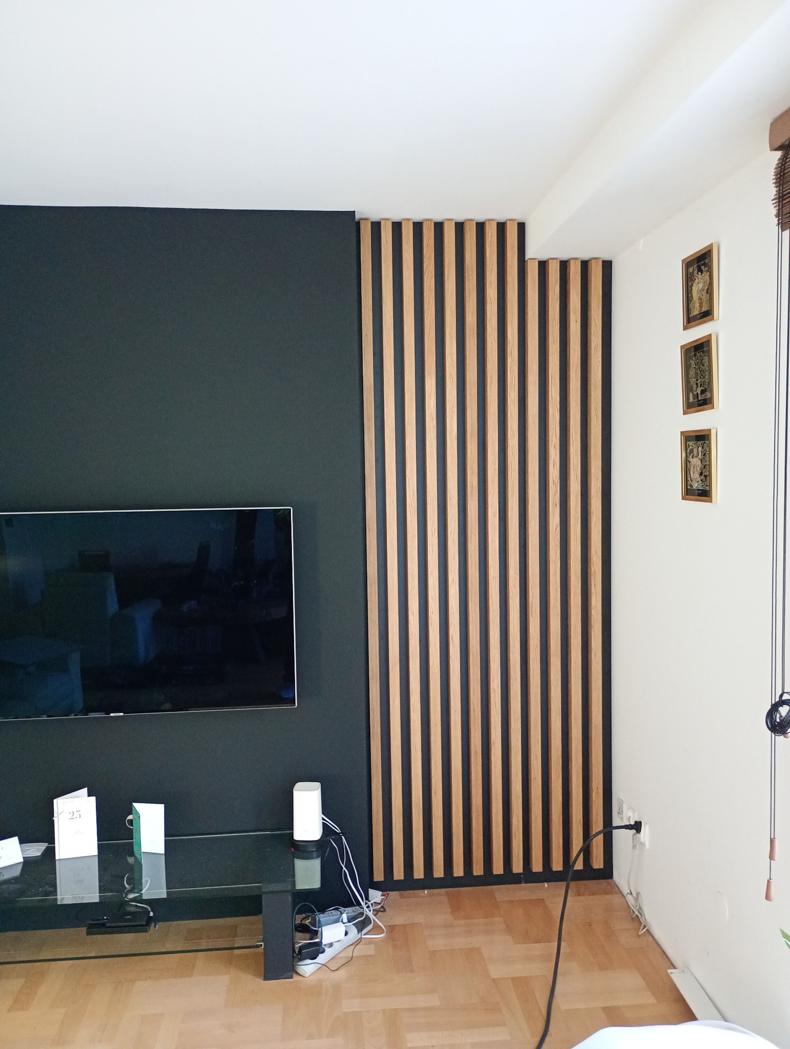 Nowoczesna aranżacja wnętrza z drewnianymi lamelami na ścianie, obok czarnej ściany z telewizorem. Widoczny fragment szklanego stolika i parkiet na podłodze.