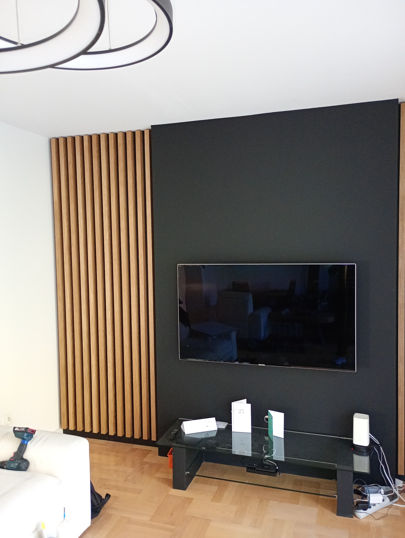 Nowoczesny salon z drewnianą ścianką działową z pionowych listew, czarną ścianą i telewizorem. Widoczny fragment białej sofy i stolik kawowy.