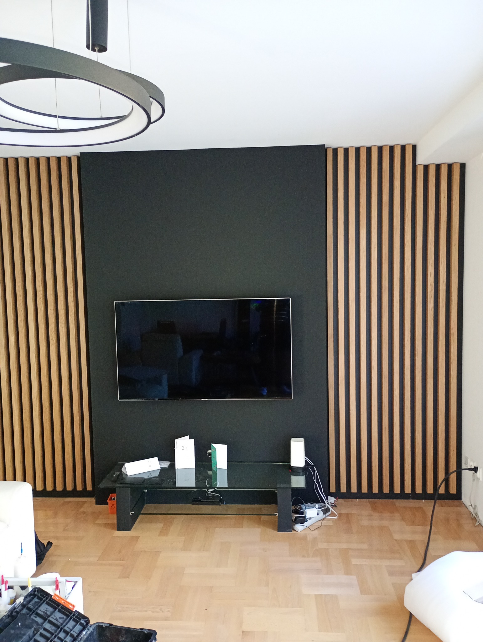 Nowoczesny salon z drewnianymi lamelami na ścianie, czarnym tłem pod telewizorem i minimalistycznym oświetleniem. Widoczny fragment podłogi z parkietu.