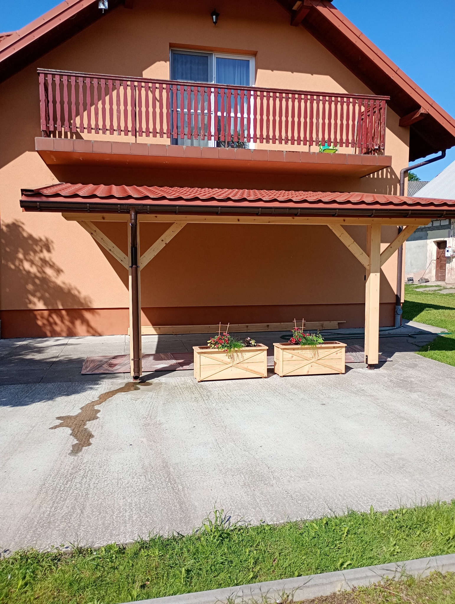 Drewniana wiata z czerwonym dachem przy domu z balkonem. Dwie drewniane donice z kwiatami. Widoczny cień i trawa. Słoneczny dzień.