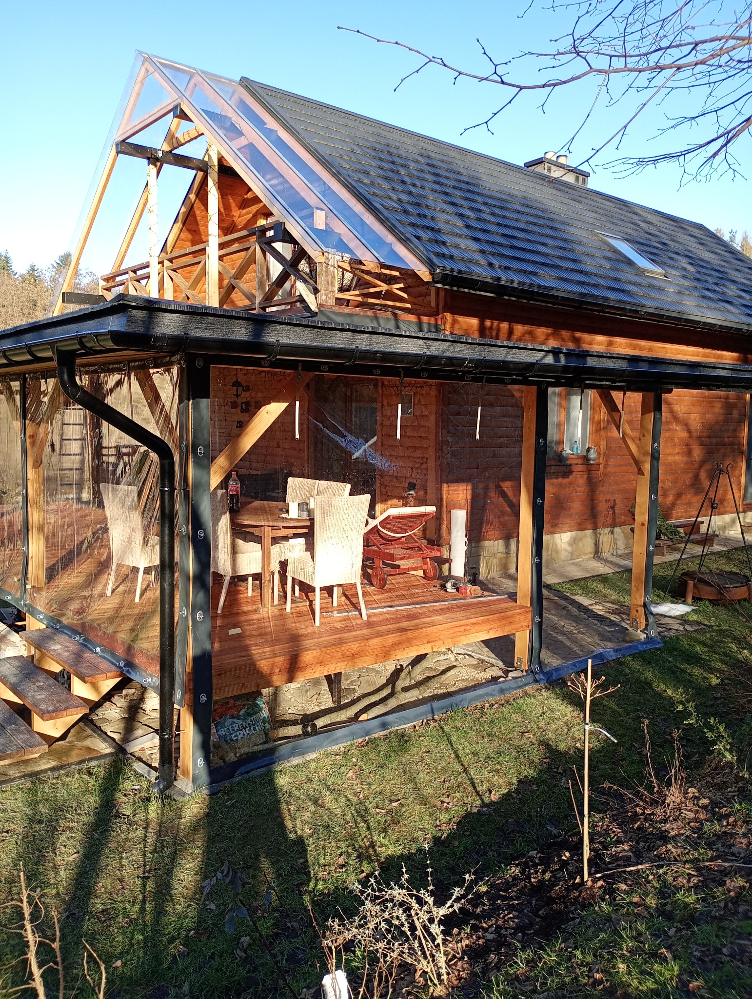 Zadaszony taras z drewna z meblami ogrodowymi i przezroczystymi osłonami bocznymi, widok z zewnątrz w słoneczny dzień. Elementy drewniane i metalowe.