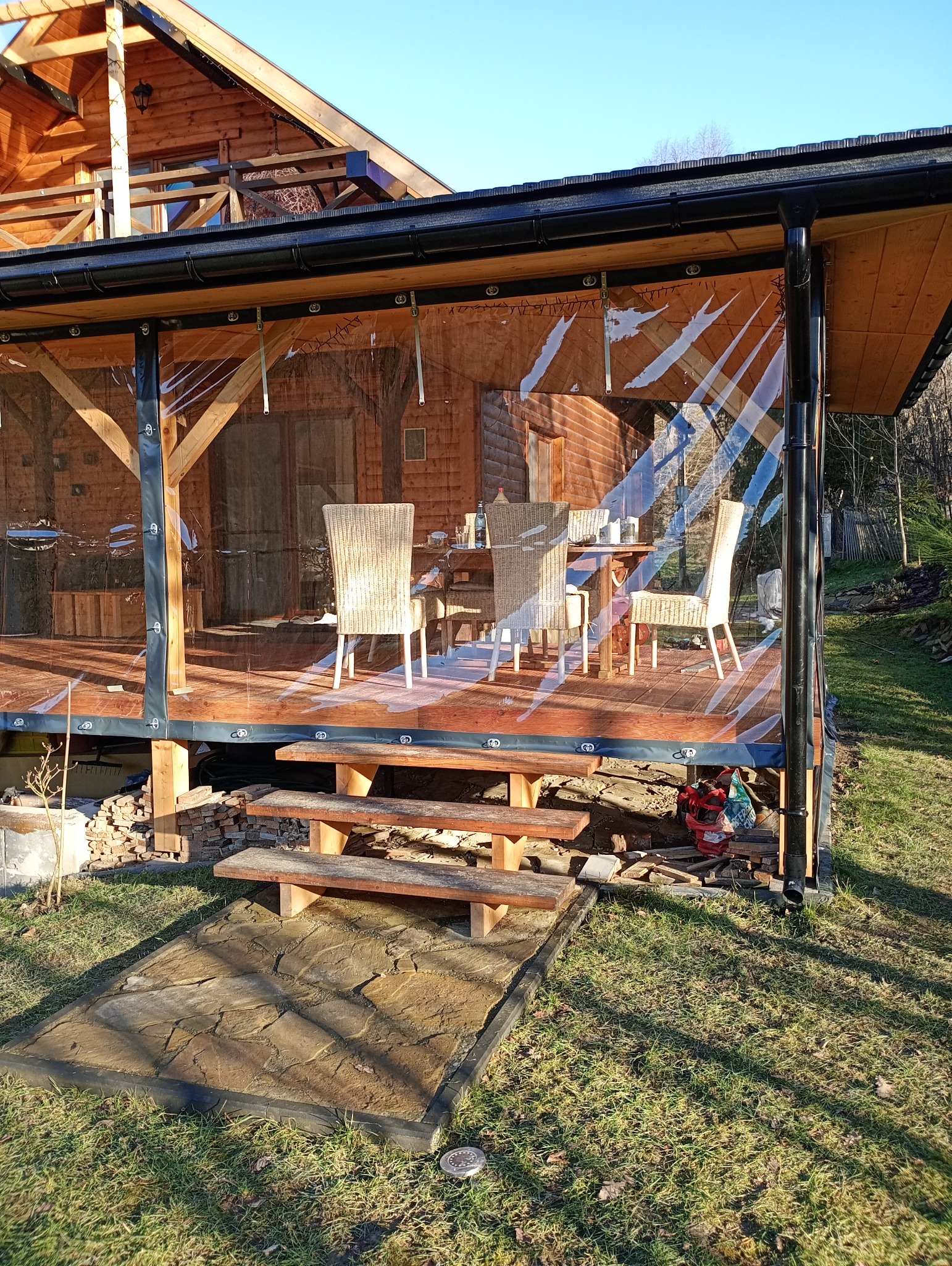 Zadaszony taras drewniany z meblami ogrodowymi i schodami wejściowymi. Przezroczyste ściany boczne chronią przed wiatrem. Widok na drewniany dom i zieleń.