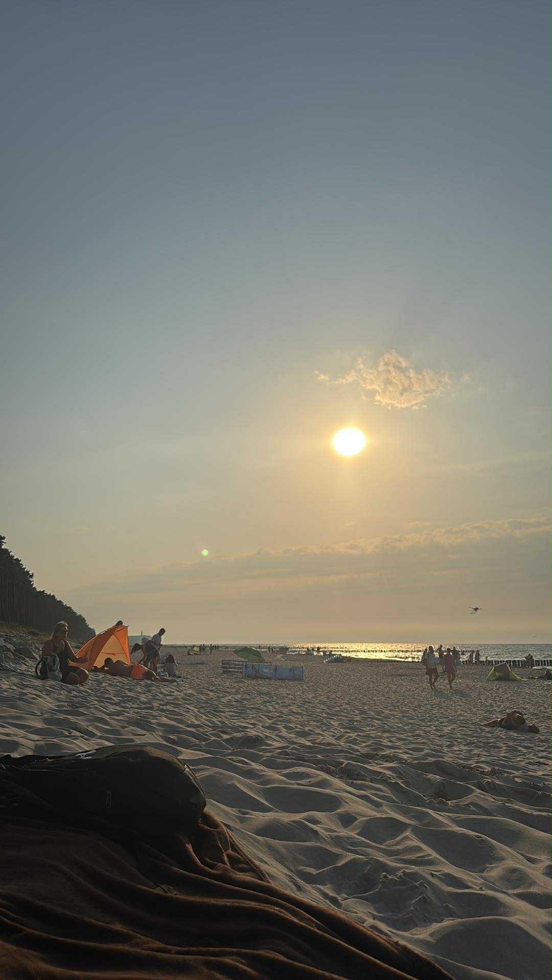 Słoneczny dzień na plaży: piasek, morze, ludzie relaksujący się pod pomarańczowym parawanem, w tle las i zachodzące słońce odbijające się w wodzie.