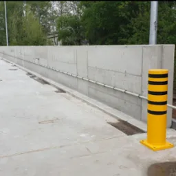 Betonowa konstrukcja z rurami i żółto-czarnym słupkiem ostrzegawczym. Widoczny fragment torów i studzienka kanalizacyjna. Projekt inżynieryjny w otoczeniu zieleni.