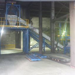 Kierownik robót instalacji sanitarnych - wykonawstwo, nadzór, projekt - Konstrukcja stalowa w hali przemysłowej: schody, platforma i przenośnik taśmowy w kolorach niebieskim i żółtym. Elementy konstrukcyjne na podłodze, surowy beton w tle.