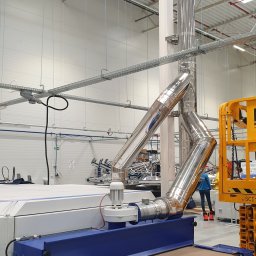 Kierownik robót instalacji sanitarnych - wykonawstwo, nadzór, projekt - Instalacja odpylająca z polerowanej stali nierdzewnej podłączona do maszyny przemysłowej w hali produkcyjnej z podnośnikiem koszowym w tle.