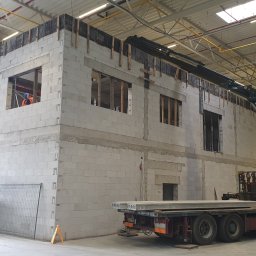 Kierownik robót instalacji sanitarnych - wykonawstwo, nadzór, projekt - Wnętrze hali w budowie: ściany z betonu komórkowego z otworami okiennymi, dźwig z boku, podnośniki koszowe i pracownik w pomarańczowym ubraniu w jednym z okien.