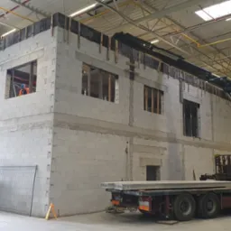 Wnętrze hali w budowie: ściany z betonu komórkowego z otworami okiennymi, dźwig z boku, podnośniki koszowe i pracownik w pomarańczowym ubraniu w jednym z okien.