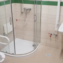 Jasna łazienka z kabiną prysznicową, umywalką i toaletą, wyposażona w uchwyty dla osób z niepełnosprawnościami. Wykończenie w beżowych i zielonych tonacjach.