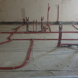 Kierownik robót instalacji sanitarnych - wykonawstwo, nadzór, projekt - Rozległa instalacja ogrzewania podłogowego z czerwonych rur, częściowo przymocowanych taśmą, na surowym podłożu w trakcie realizacji. Widoczne pionowe wyprowadzenia.