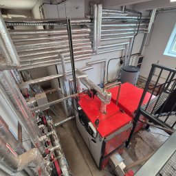 Kierownik robót instalacji sanitarnych - wykonawstwo, nadzór, projekt - Wysoki kąt: Kotłownia z czerwonym kotłem i licznymi izolowanymi rurami. Widoczne elementy instalacji, drabina i okno z widokiem na zieleń.