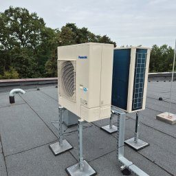 Kierownik robót instalacji sanitarnych - wykonawstwo, nadzór, projekt - Dwa klimatyzatory Panasonic na dachu pokrytym papą, zamontowane na metalowych konstrukcjach. Widoczne drzewa w tle i pochmurne niebo.