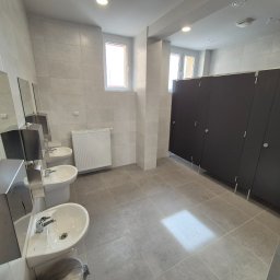 Kierownik robót instalacji sanitarnych - wykonawstwo, nadzór, projekt - Wykończona toaleta publiczna z trzema umywalkami, lustrami, grzejnikiem i kabinami WC. Wnętrze wyłożone szarymi płytkami. Widoczne detale armatury i oświetlenia.