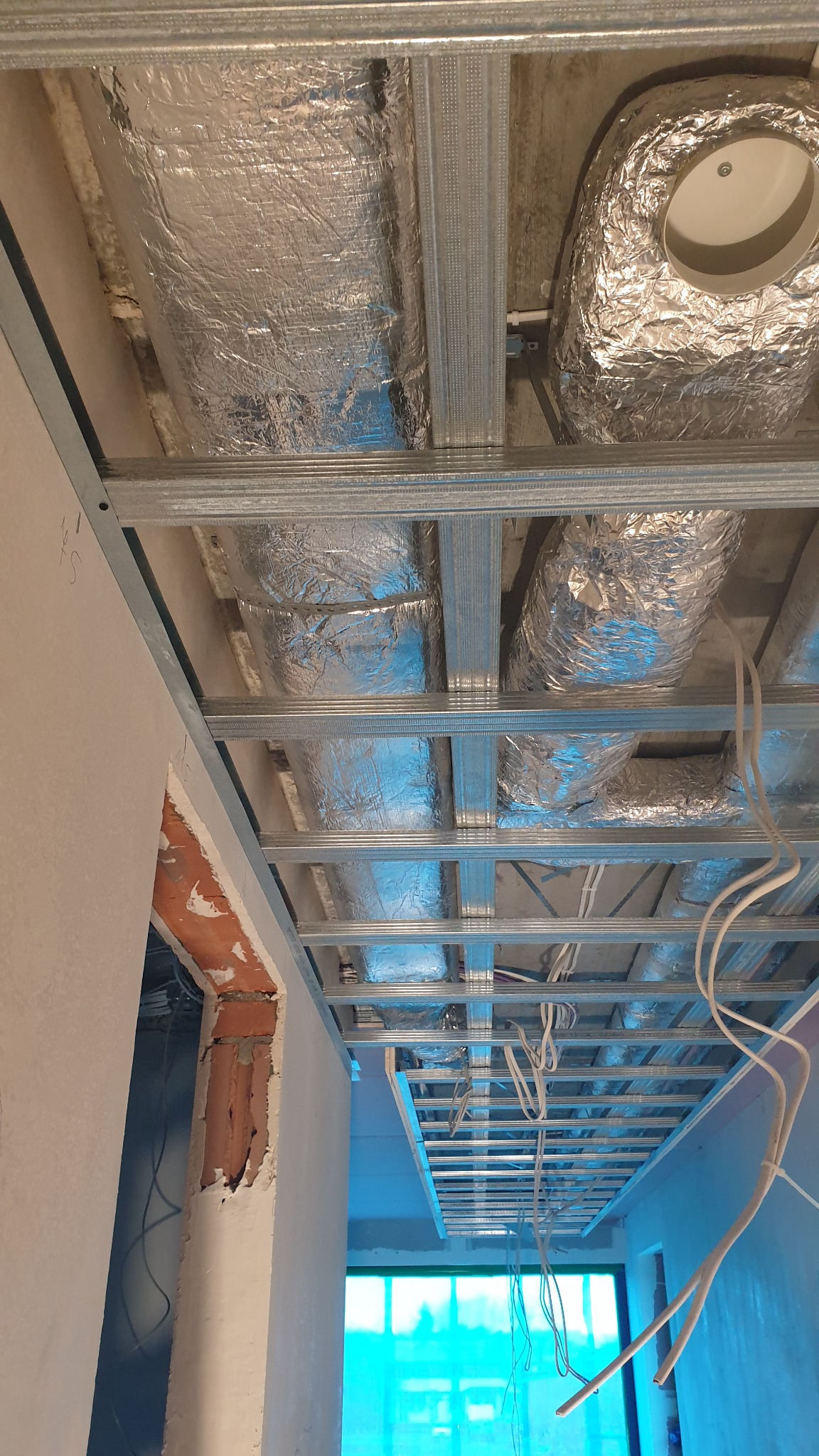 Srebrne rury wentylacyjne pod sufitem podwieszanym z metalową konstrukcją. Widoczne kable i fragment ściany z cegłą. Ujęcie z dołu, w trakcie montażu.