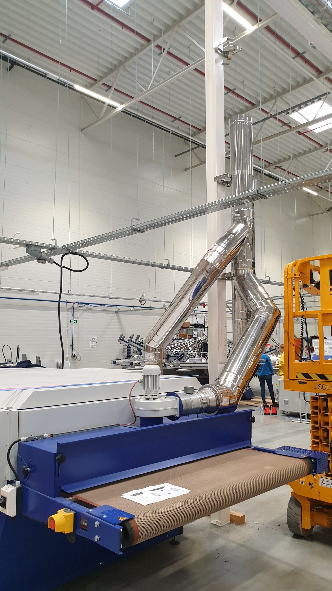 Instalacja odpylająca z polerowanej stali nierdzewnej podłączona do maszyny przemysłowej w hali produkcyjnej z podnośnikiem koszowym w tle.