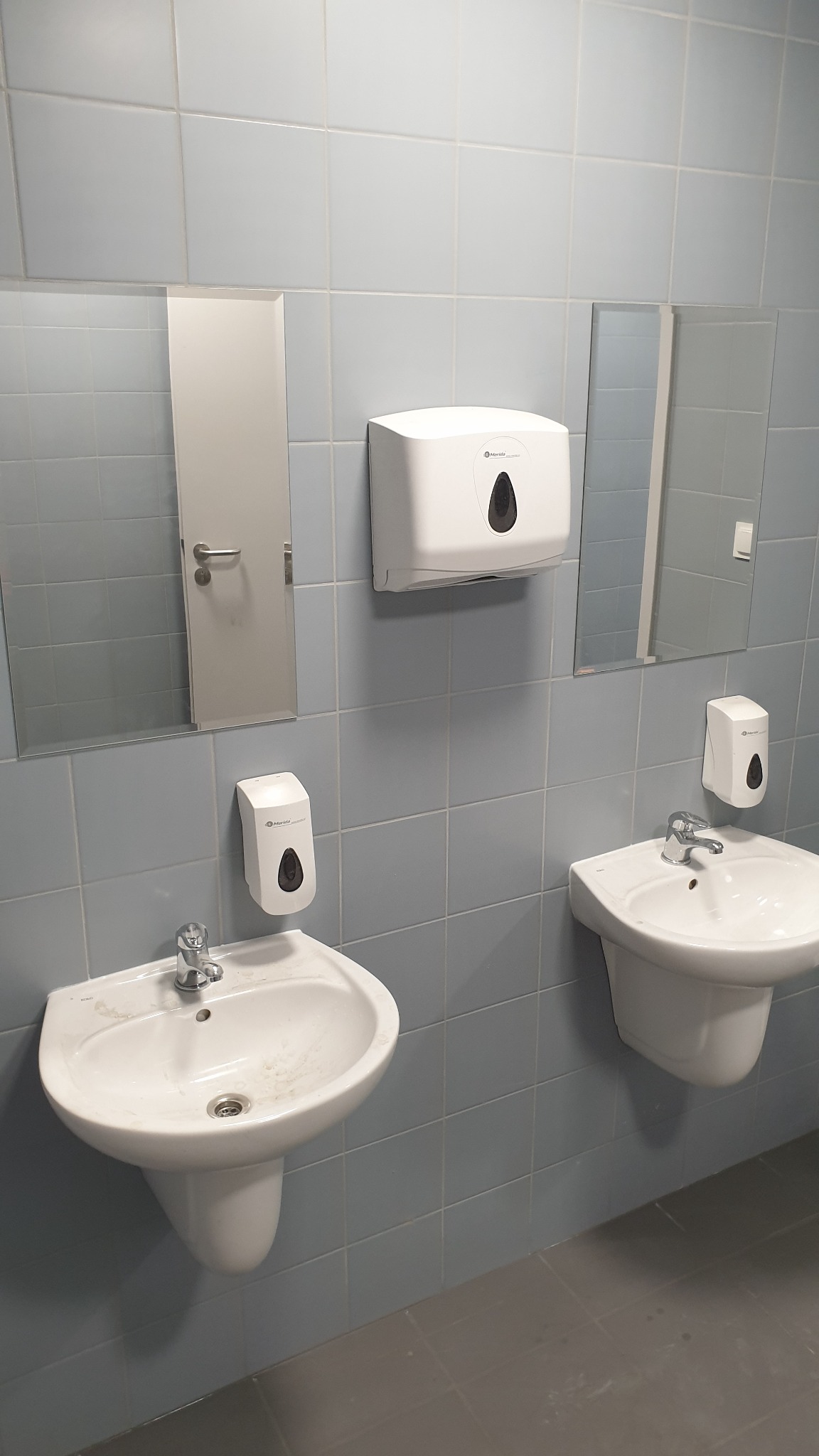 Umywalki z armaturą i dozownikami mydła w pomieszczeniu wyłożonym jasnoniebieskimi płytkami. Widoczne lustra i podajnik ręczników. Ujęcie z poziomu podłogi.