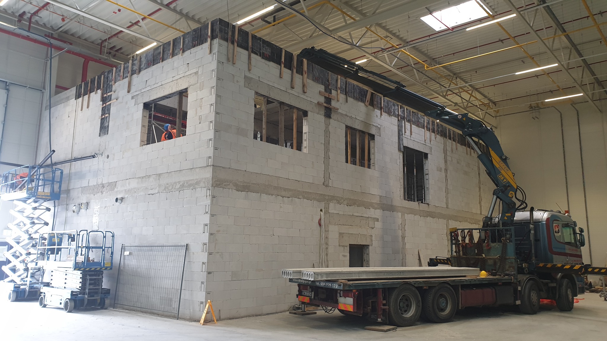 Wnętrze hali w budowie: ściany z betonu komórkowego z otworami okiennymi, dźwig z boku, podnośniki koszowe i pracownik w pomarańczowym ubraniu w jednym z okien.