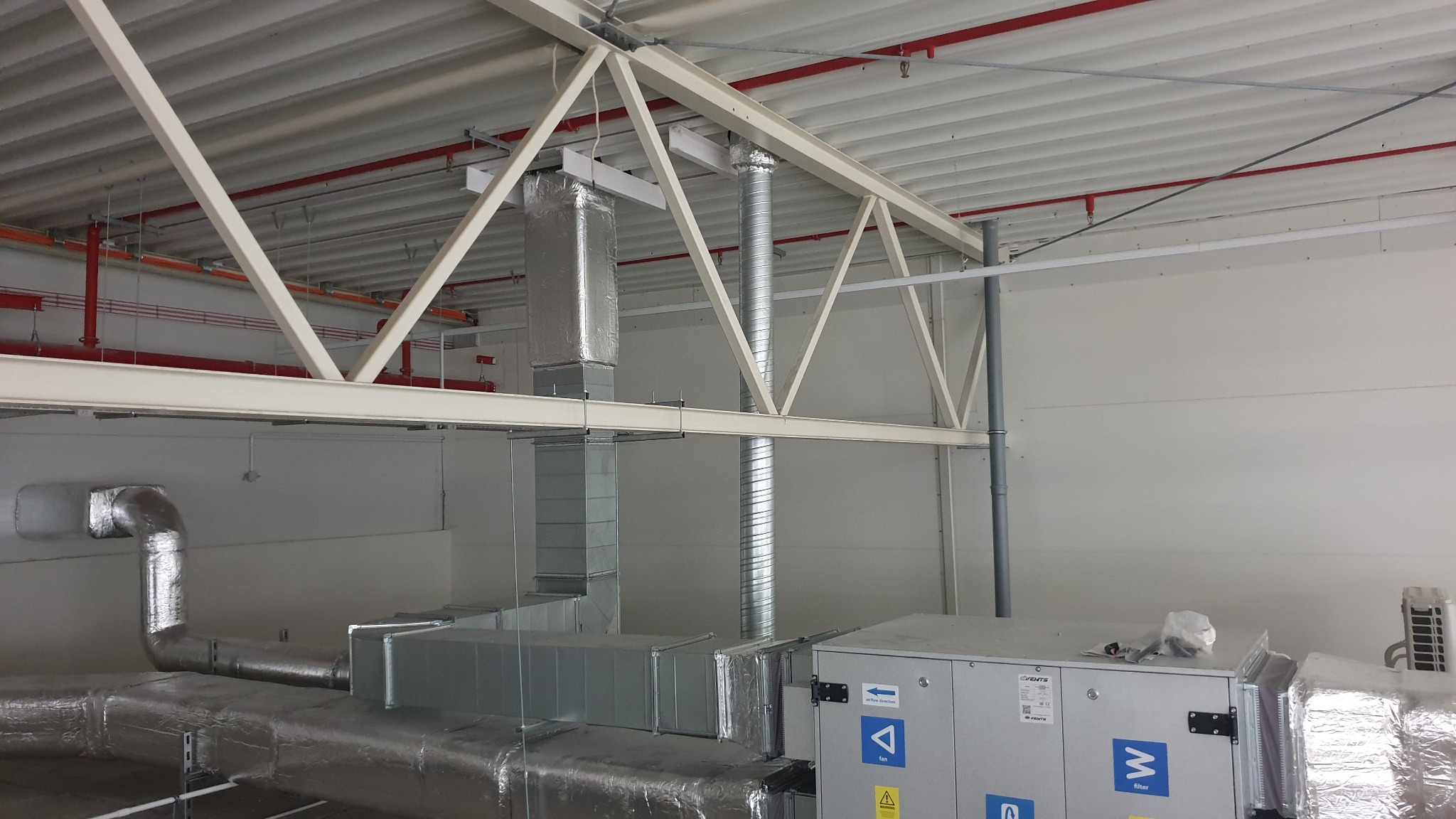Instalacja wentylacyjna w hali: srebrne rury i izolacja, widoczne elementy konstrukcji stalowej, szare ściany. Detal systemu wentylacji w budynku przemysłowym.