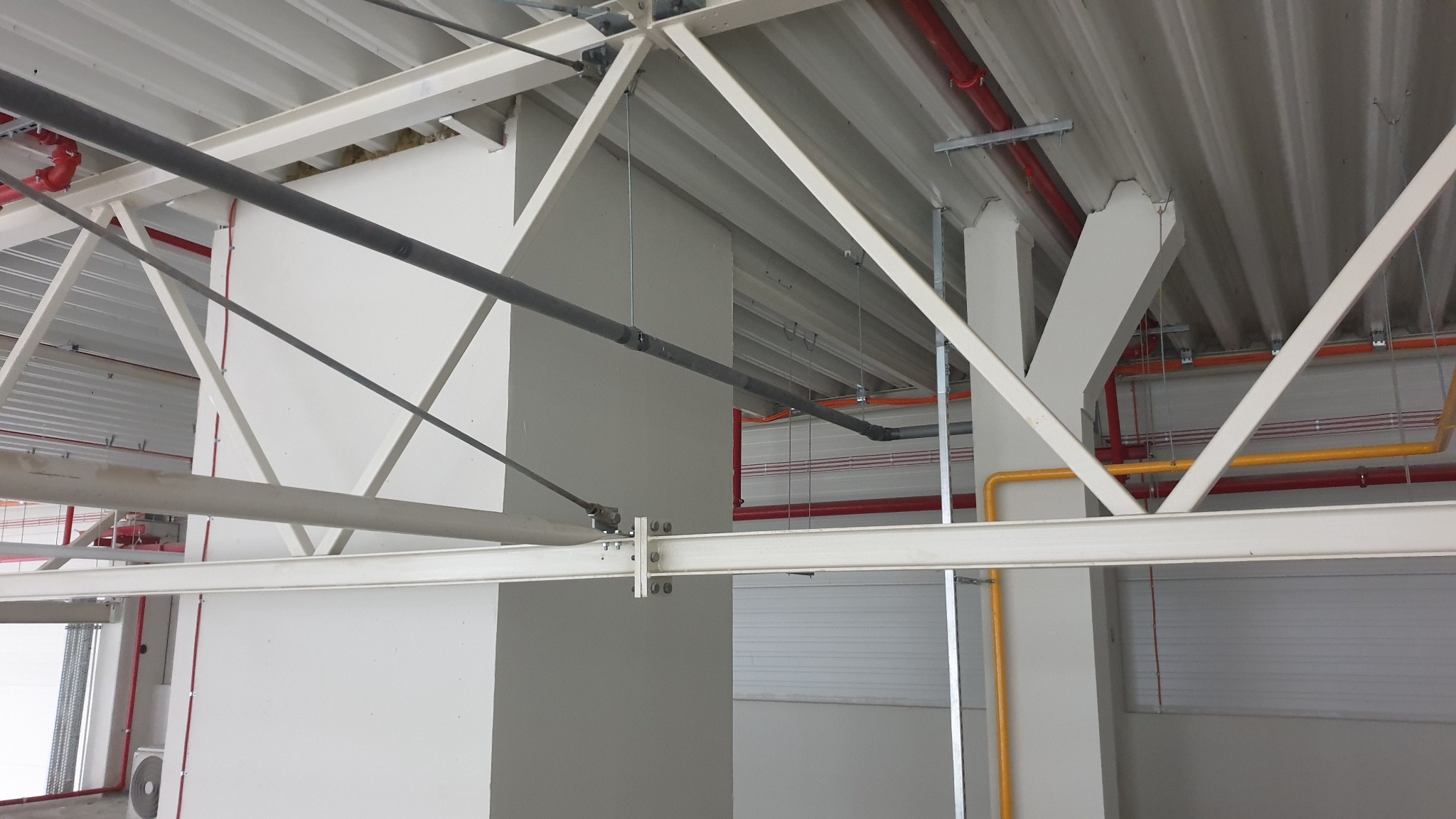 Instalacje w hali: rury w kolorach czerwonym, żółtym i szarym, biegnące pod sufitem z blachy trapezowej, wsparte na stalowej konstrukcji nośnej.