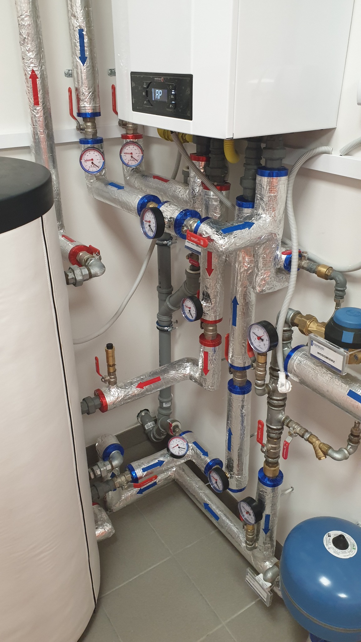 Złożona instalacja grzewcza z kotłem, rurami z izolacją, manometrami i zaworami, umieszczona w pomieszczeniu z szarą podłogą.