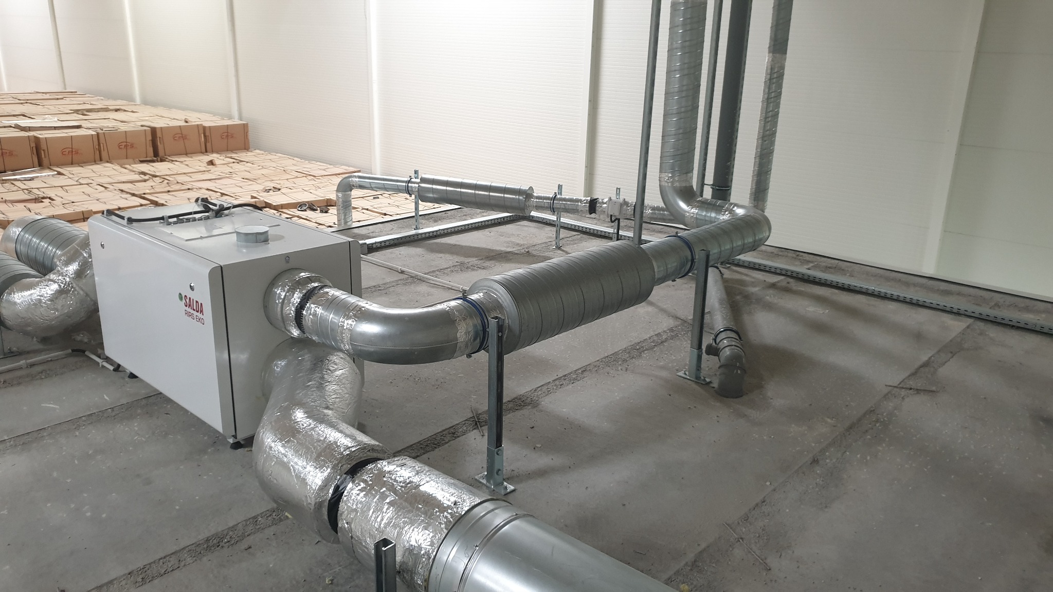 Szara centrala wentylacyjna Salda RIRS EKO z podłączonymi, izolowanymi przewodami wentylacyjnymi w hali. Widoczne podwieszenia i mocowania instalacji.