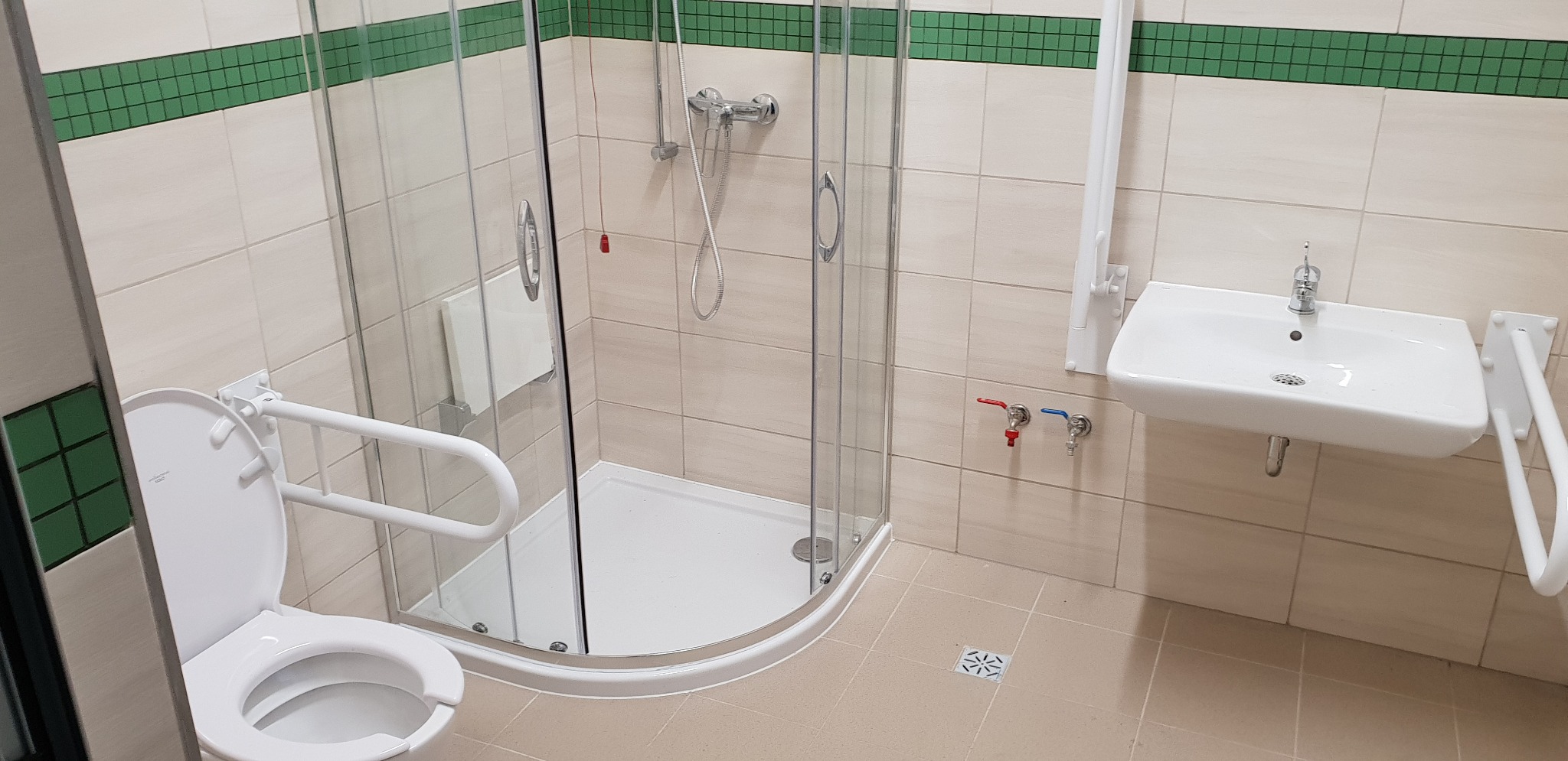 Jasna łazienka z kabiną prysznicową, umywalką i toaletą, wyposażona w uchwyty dla osób z niepełnosprawnościami. Wykończenie w beżowych i zielonych tonacjach.