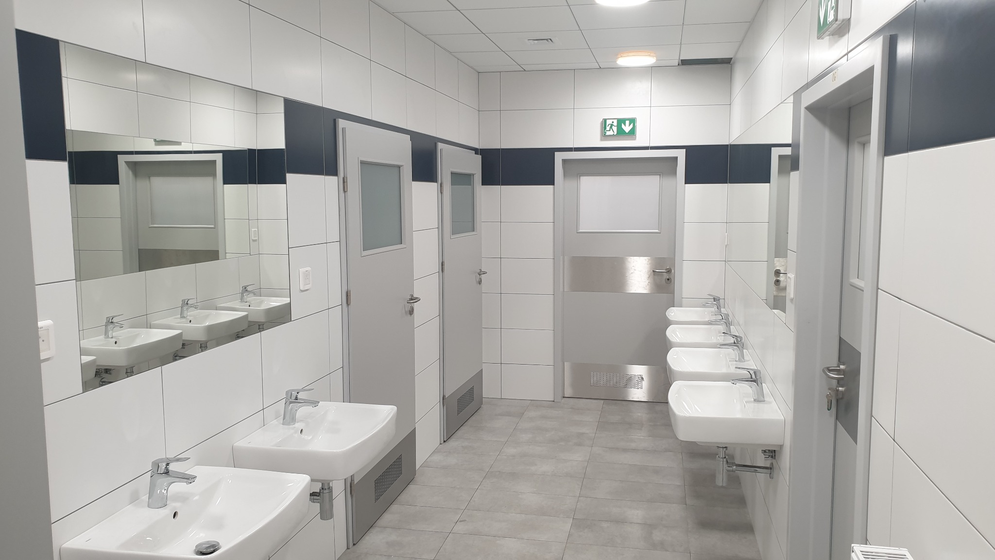 Wnętrze publicznej toalety z rzędem umywalek i lustrem, białe płytki z szarym pasem, szare drzwi z oznaczeniem ewakuacyjnym. Minimalistyczny design.