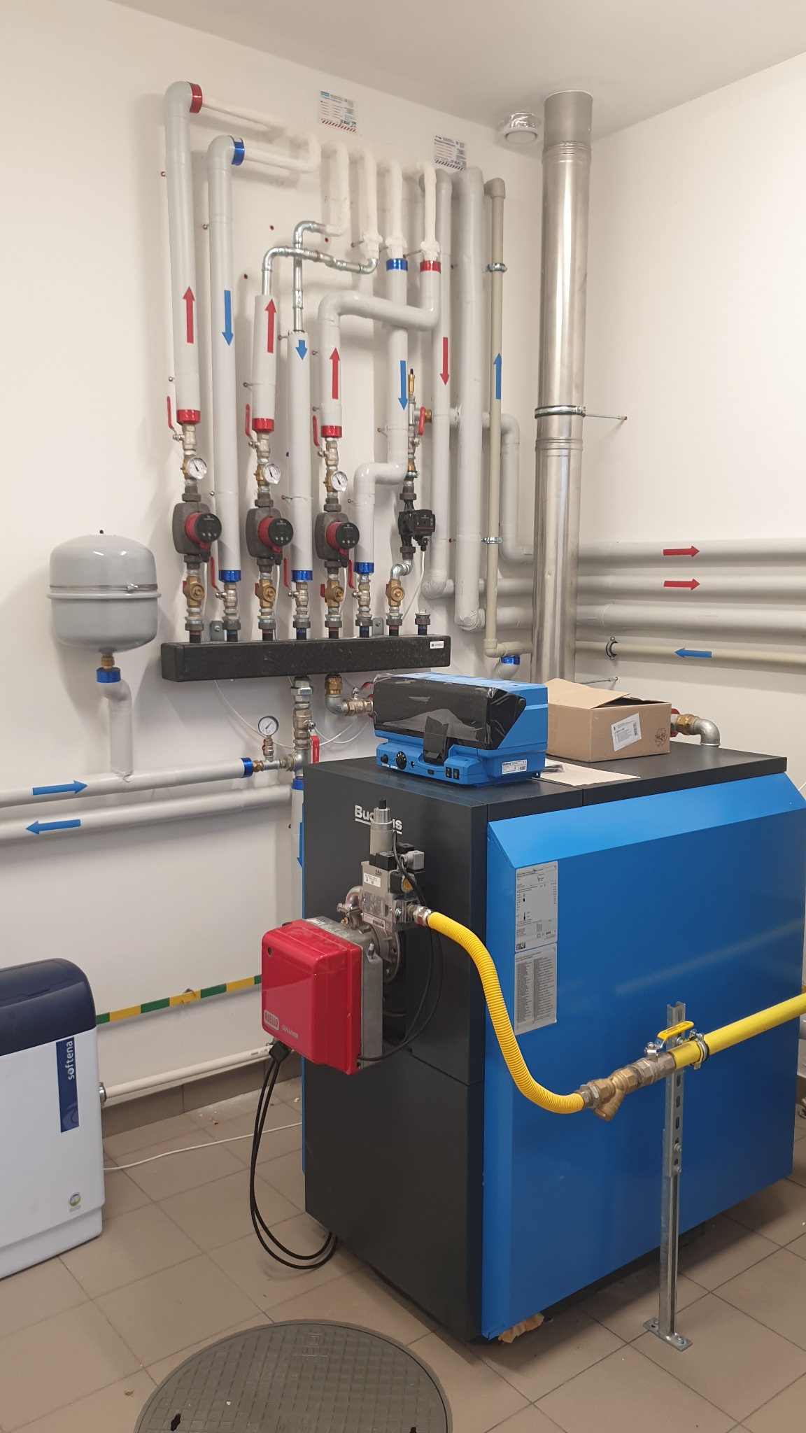 Kotłownia z nowoczesnym kotłem grzewczym Buderus, systemem rur z oznaczeniami przepływu i elementami sterującymi. Widoczne detale instalacji i podłączenia.