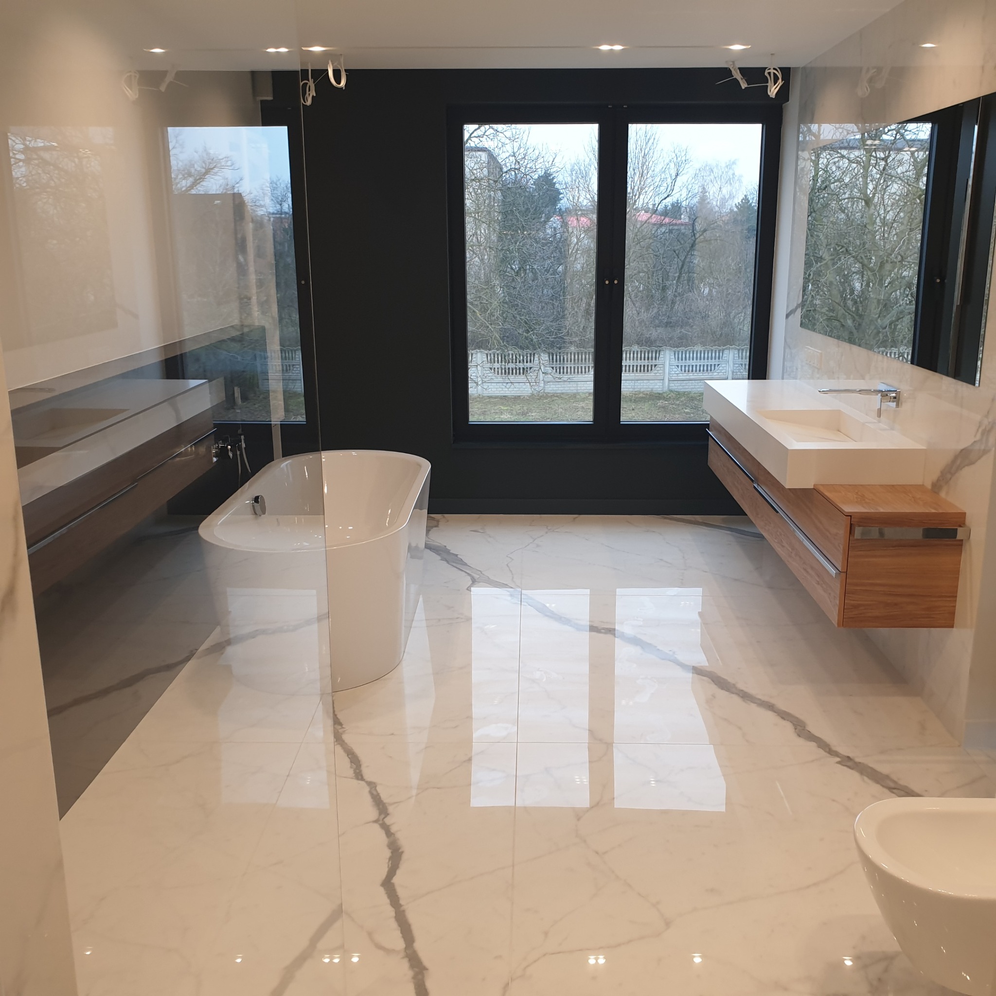 Nowoczesna łazienka z marmurową podłogą, wolnostojącą wanną i wiszącą szafką z umywalką. Duże okno z widokiem na drzewa. Minimalistyczny design.