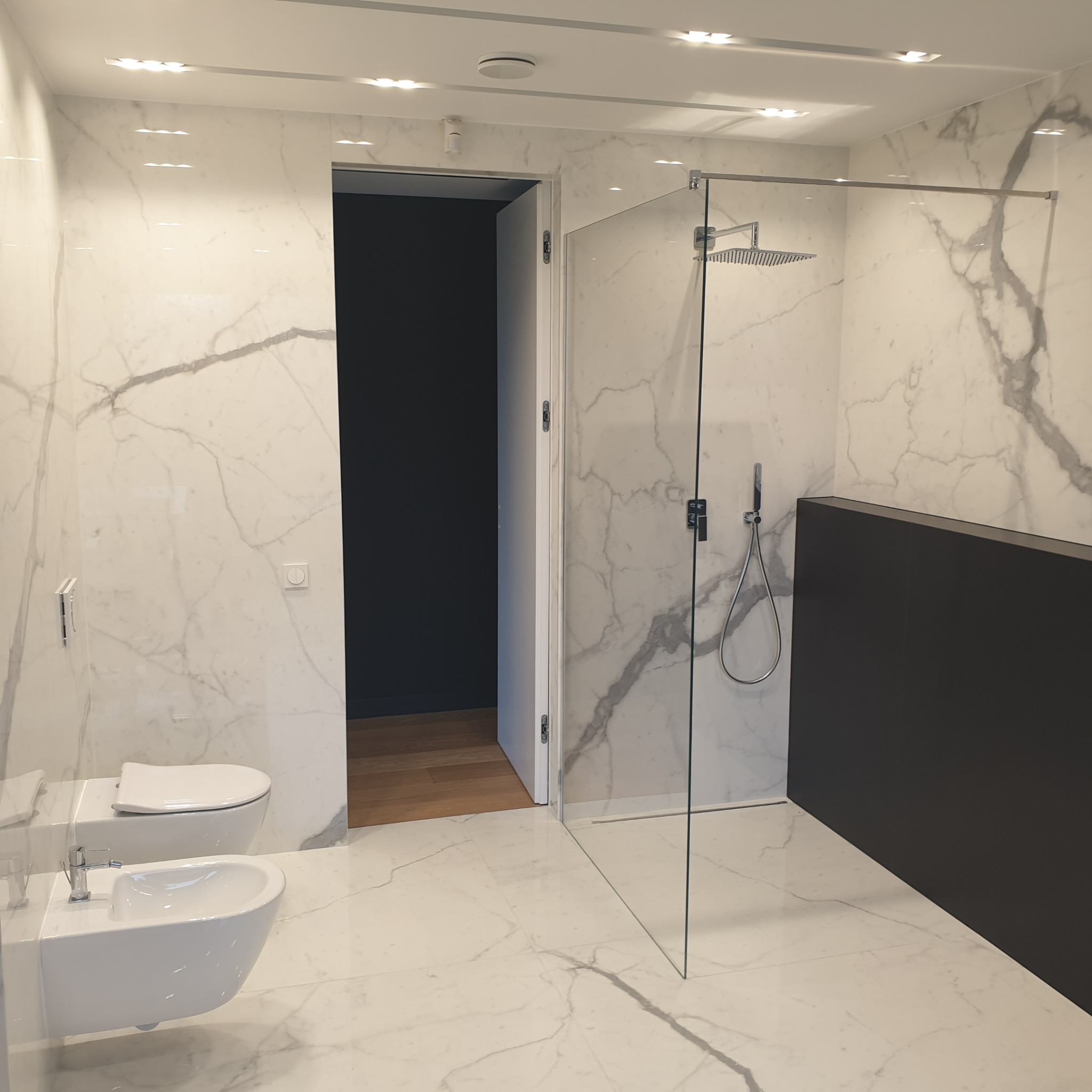 Nowoczesna łazienka z kabiną prysznicową typu walk-in, wykończona białym marmurem z szarymi żyłami. Widoczne białe WC i bidet podwieszane. Oświetlenie sufitowe.