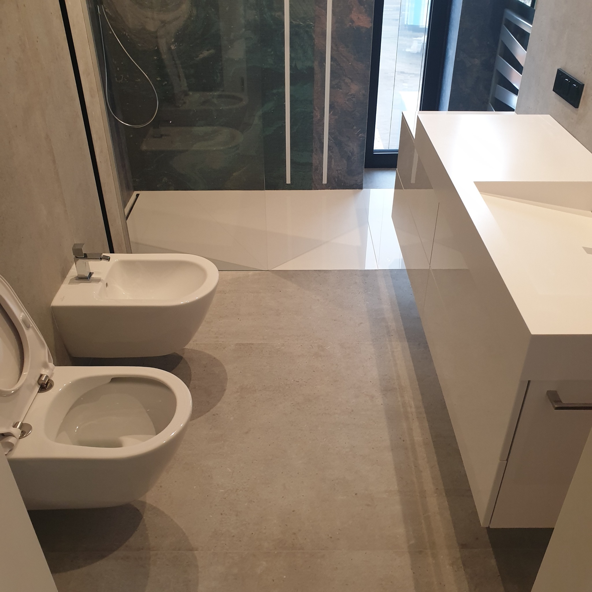 Nowoczesna łazienka z białymi sanitariatami: toaleta z otwartą klapą, bidet i szafka pod umywalkę. Minimalistyczny design, szare płytki podłogowe i szklana kabina prysznicowa.
