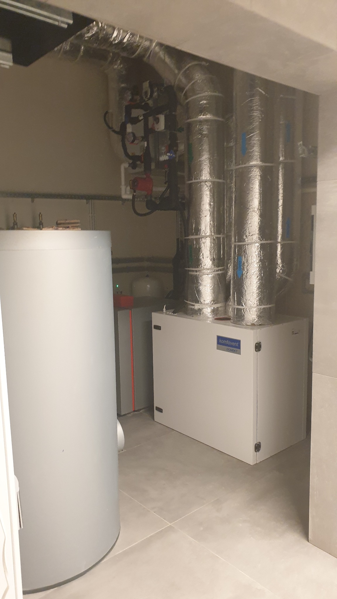 Kotłownia z białym bojlerem, szarym piecem i systemem rur z izolacją. Widoczne także elementy instalacji grzewczej i wentylacyjnej.