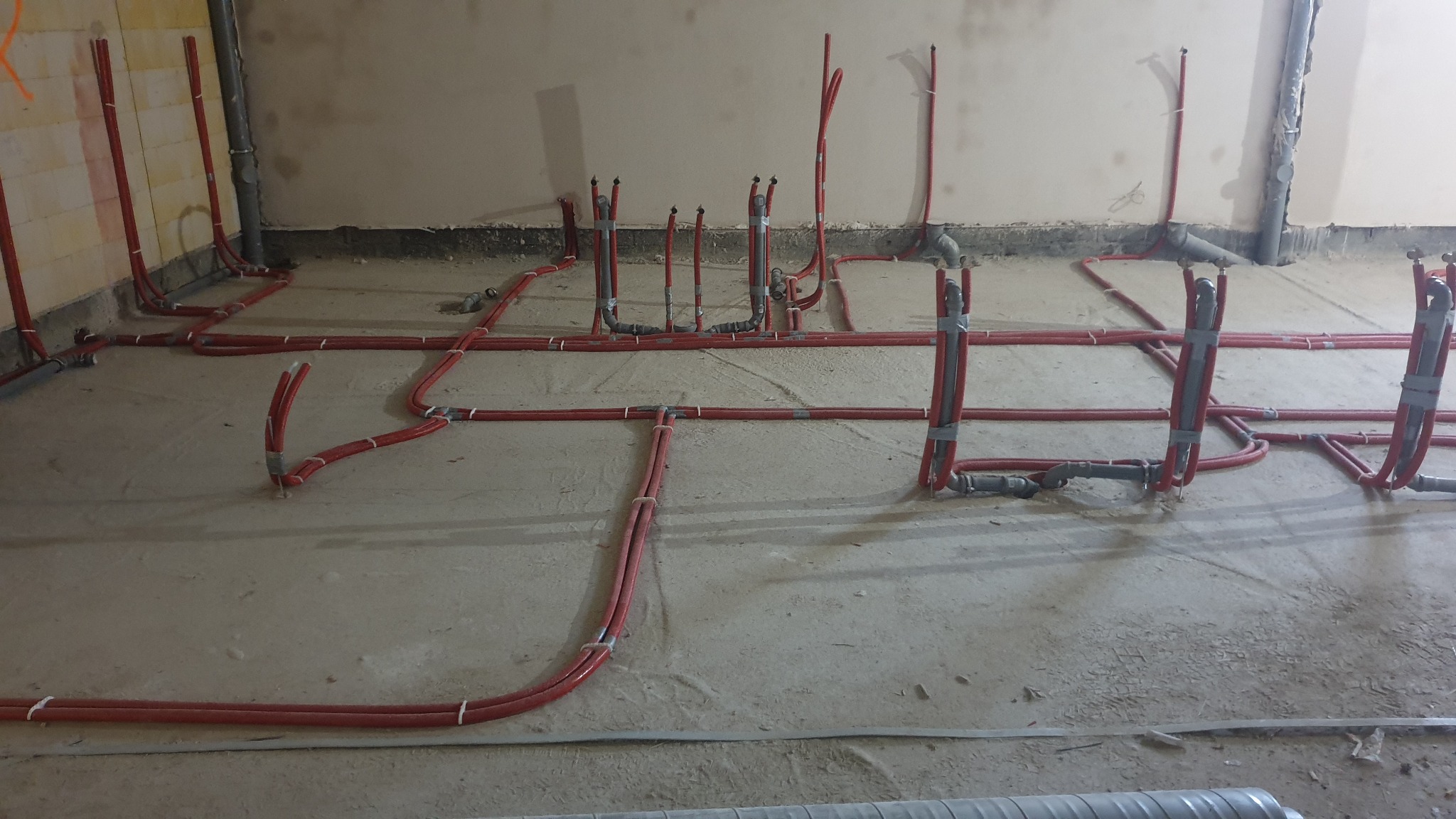 Rozległa instalacja ogrzewania podłogowego z czerwonych rur, częściowo przymocowanych taśmą, na surowym podłożu w trakcie realizacji. Widoczne pionowe wyprowadzenia.