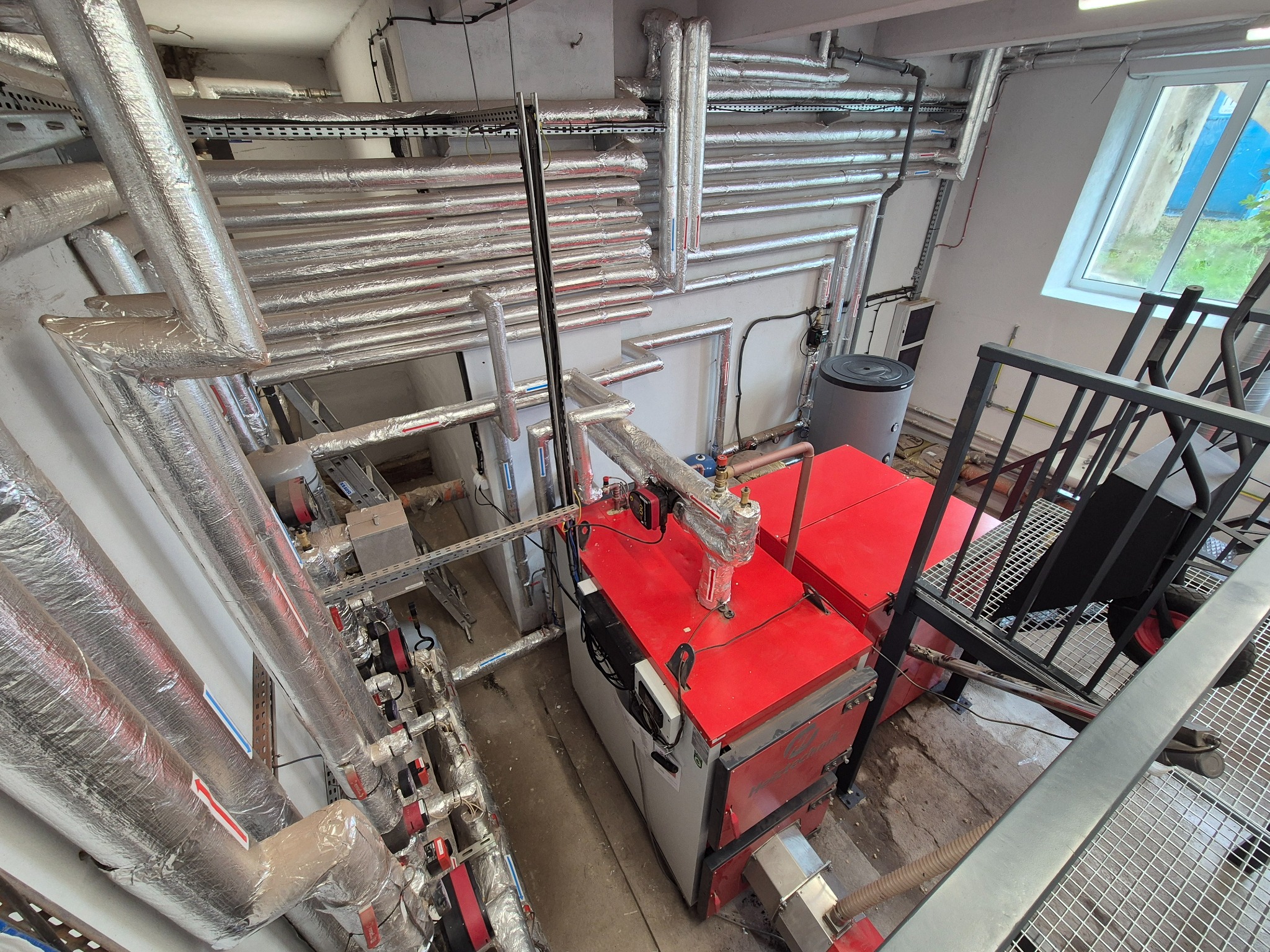 Wysoki kąt: Kotłownia z czerwonym kotłem i licznymi izolowanymi rurami. Widoczne elementy instalacji, drabina i okno z widokiem na zieleń.