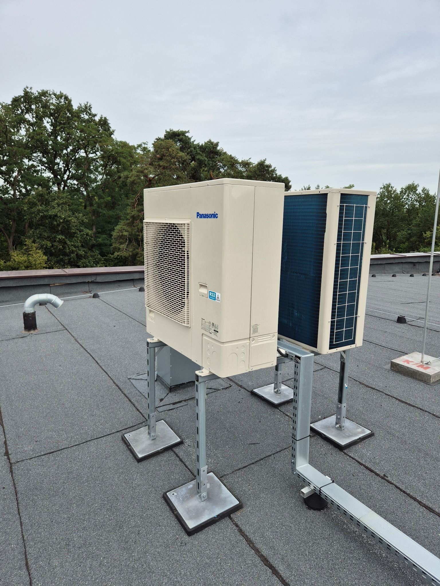Dwa klimatyzatory Panasonic na dachu pokrytym papą, zamontowane na metalowych konstrukcjach. Widoczne drzewa w tle i pochmurne niebo.