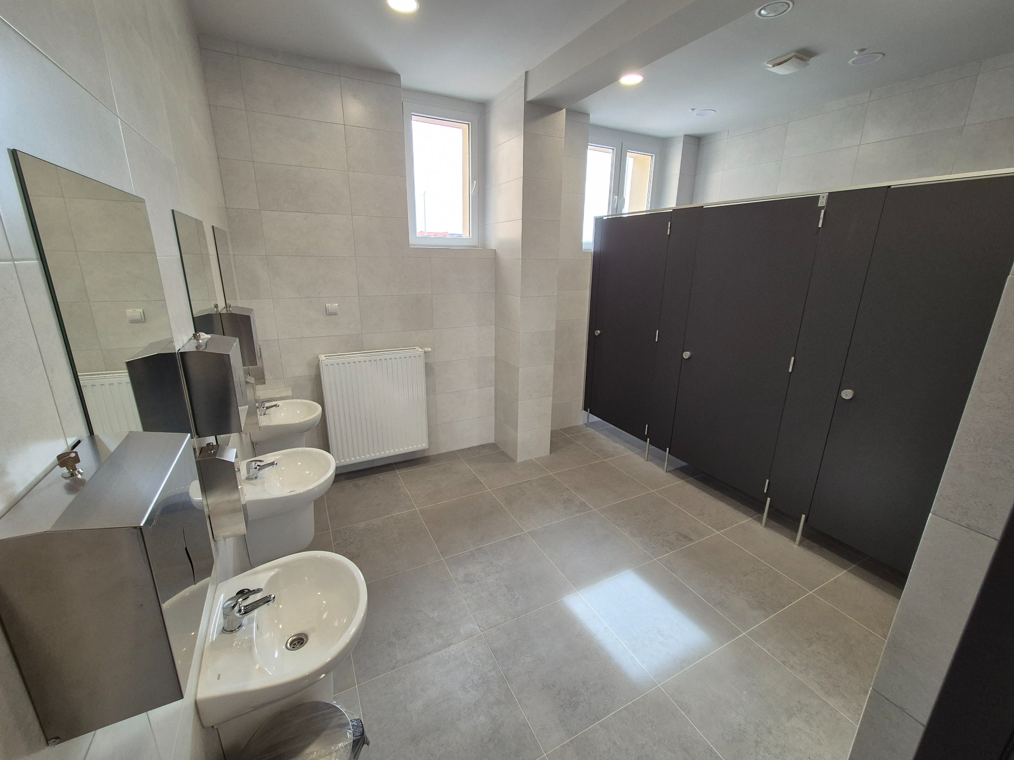 Wykończona toaleta publiczna z trzema umywalkami, lustrami, grzejnikiem i kabinami WC. Wnętrze wyłożone szarymi płytkami. Widoczne detale armatury i oświetlenia.