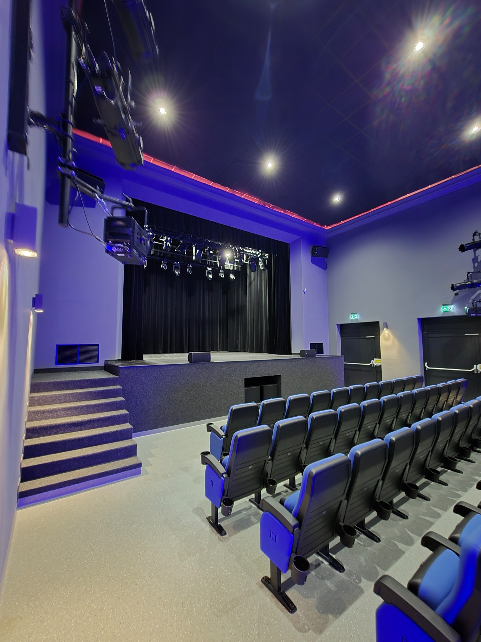 Widok sali widowiskowej w Tomaszowie Mazowieckim z rzędami niebieskich foteli i czarną sceną oświetloną reflektorami. Nowoczesne, minimalistyczne wnętrze.