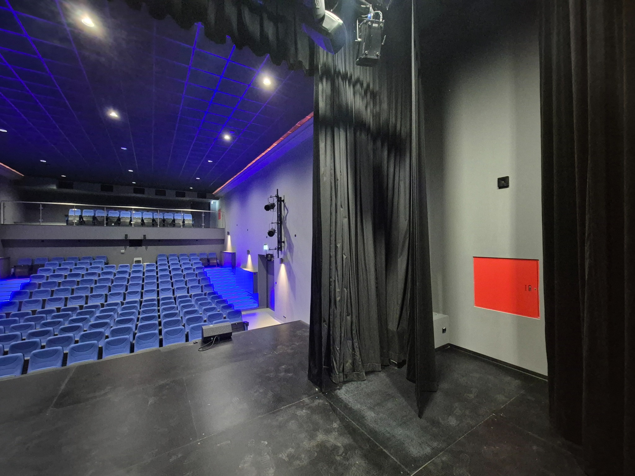Widok na salę teatralną zza kulis: scena, rzędy niebieskich foteli i oświetlenie. Ciemna kurtyna częściowo zasłania widok, dodając tajemniczości. Widoczna instalacja oświetleniowa.