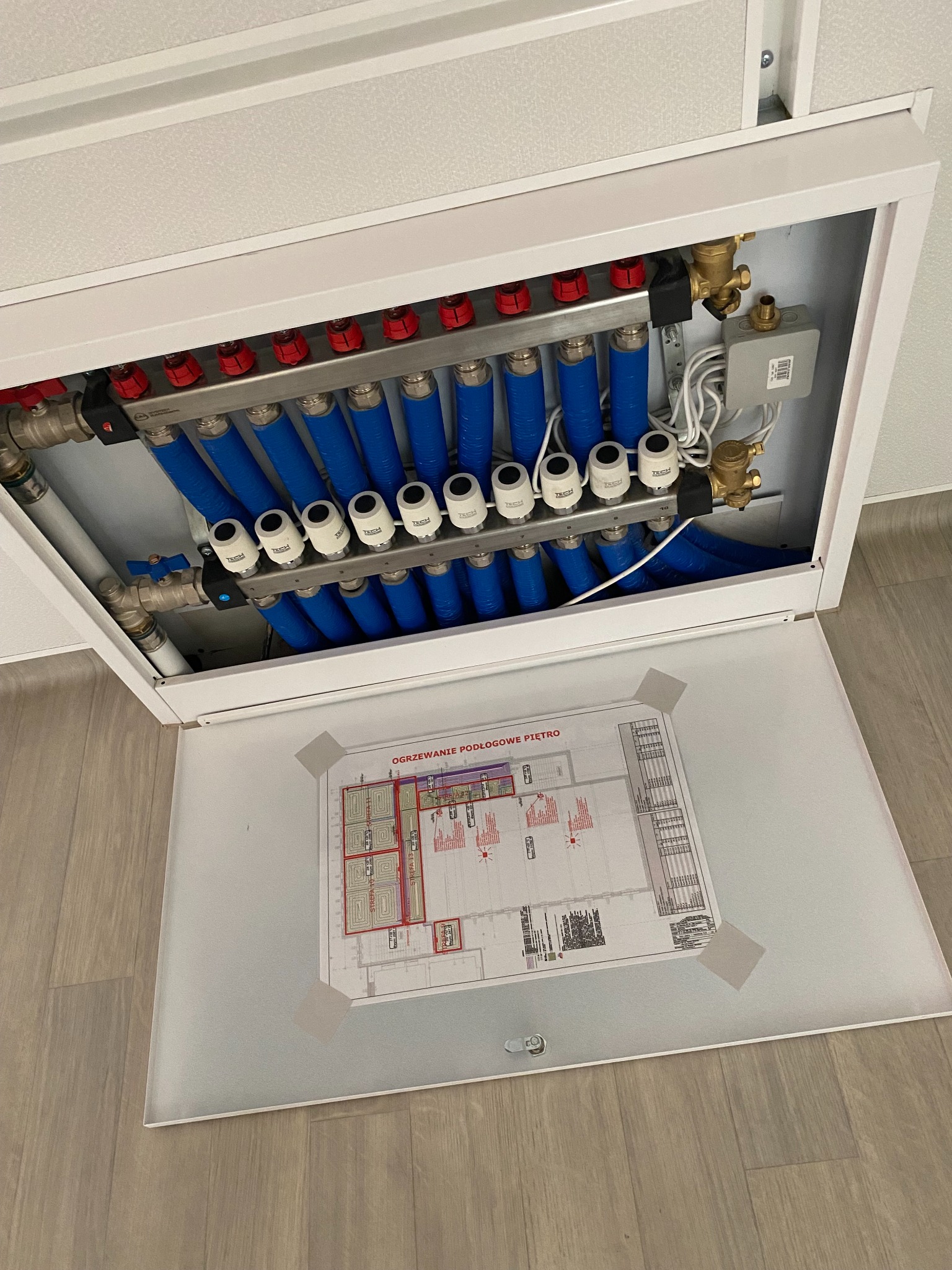 Rozdzielacz ogrzewania podłogowego w szafce, z widocznym schematem instalacji na drzwiach. Niebieskie rury i białe regulatory. Widok z góry.