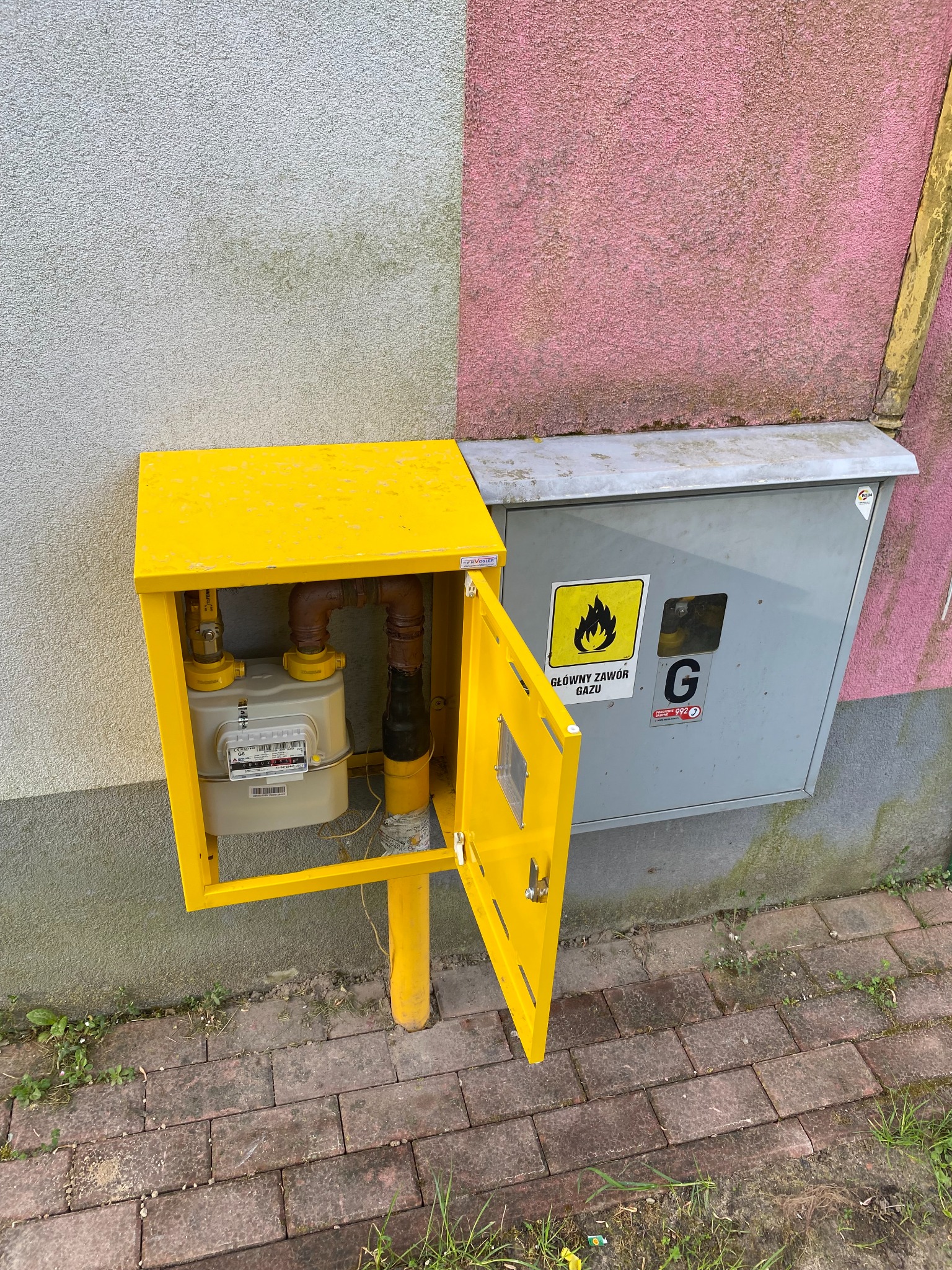 Żółta skrzynka gazowa z otwartymi drzwiczkami, obok szara skrzynka z napisem 'Główny zawór gazu', na tle ściany dwukolorowej i kostki brukowej.