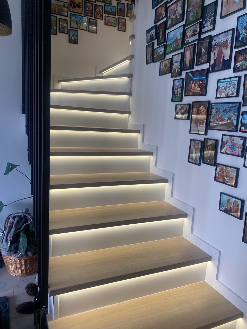 Nowoczesne schody z podświetleniem LED, drewniane stopnie i czarna balustrada, ściana udekorowana ramkami ze zdjęciami.