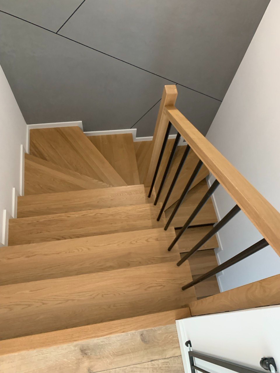 Kątowe ujęcie na drewniane schody z czarną balustradą, prowadzące w dół. Szare panele sufitowe z czarnymi liniami dodają nowoczesnego charakteru.