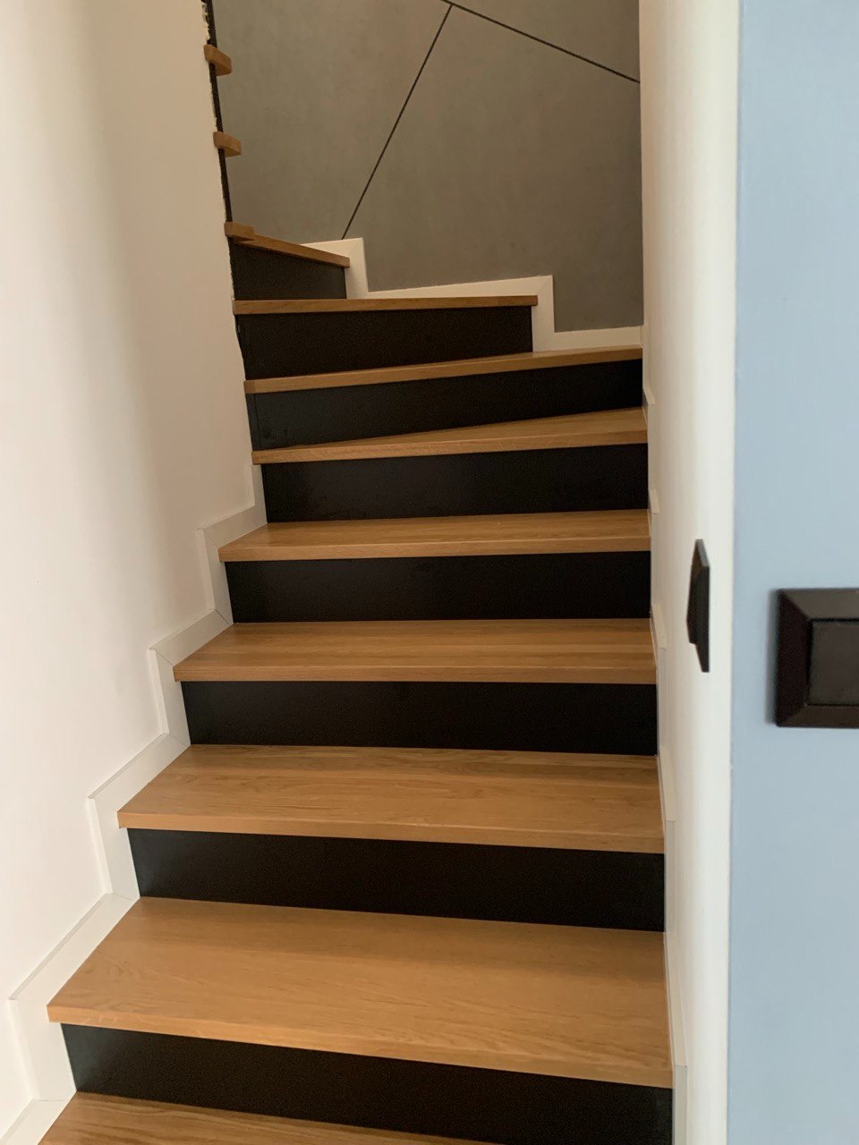 Nowoczesne schody drewniane z czarnymi podstopnicami, oświetlone naturalnym światłem, widok z dołu, minimalistyczny design, białe ściany i szara ściana w tle.