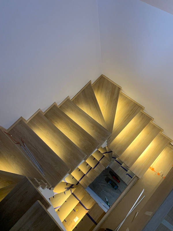 Kaskada drewnianych schodów z podświetleniem LED, widok z góry. Etap montażu, zabezpieczone folią. Jasne drewno, ciepłe światło.