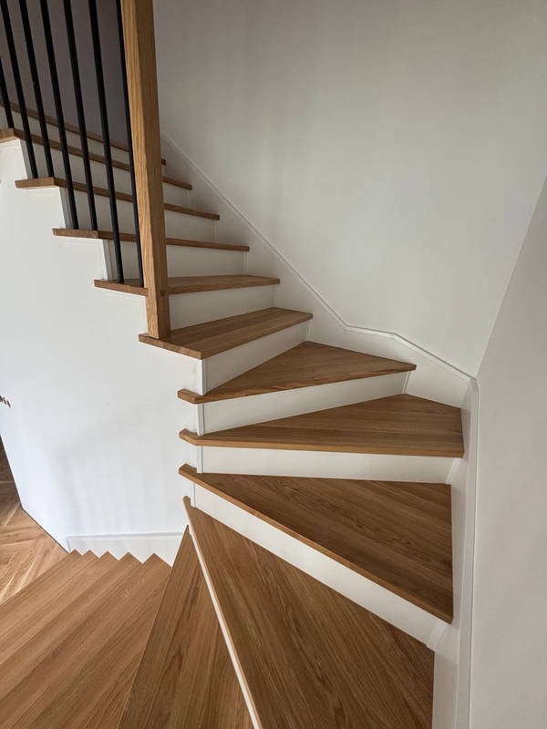 Nowoczesne schody z drewnianymi stopniami i czarną balustradą, widok z góry. Jasne wnętrze, białe ściany. Charakterystyczny wzór parkietu na podeście.